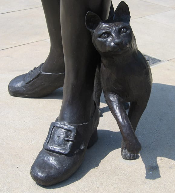 Bronze statue of a cat rubbing against a person's legs with buckled shoes.