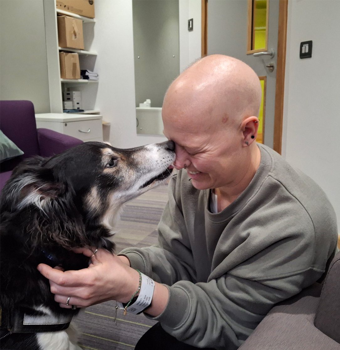 Bald person with hospital band smiling, nose-to-nose with a dog they're holding.