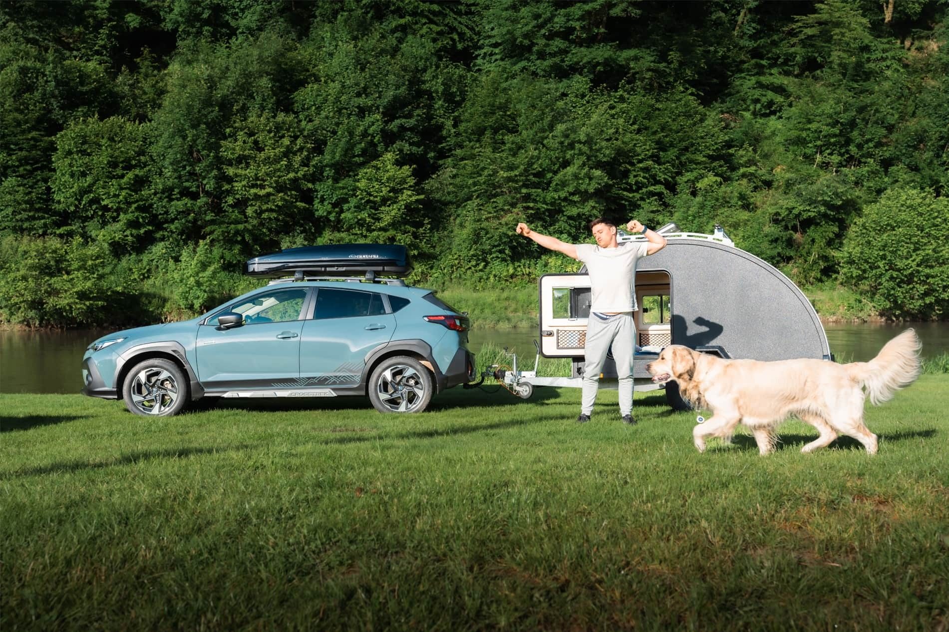 Blue Subaru Crosstrek towing teardrop trailer, man stretching, golden retriever walking nearby.