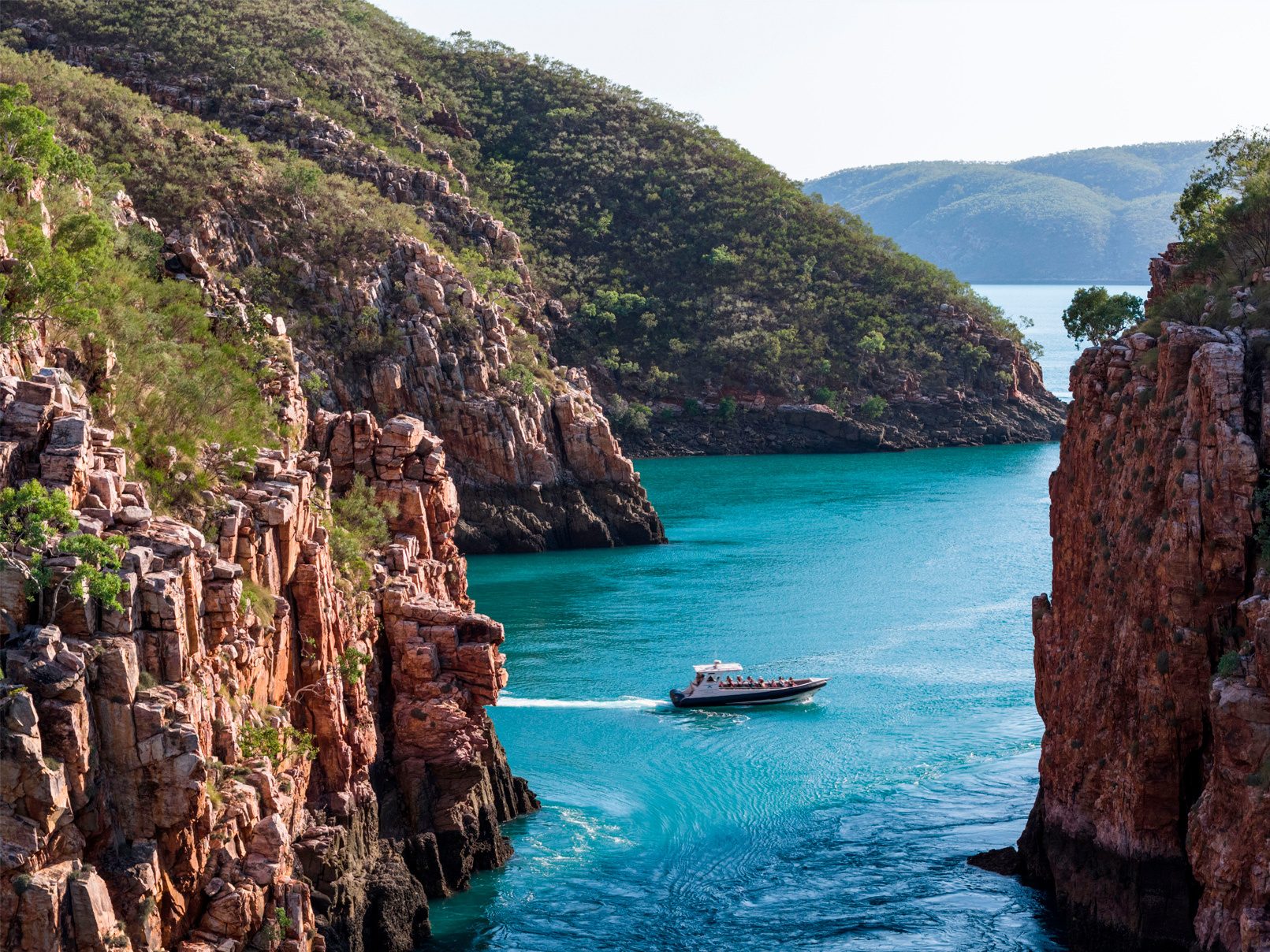 A powerboat speeds through a narrow turquoise channel between steep, rugged cliffs covered with green vegetation.