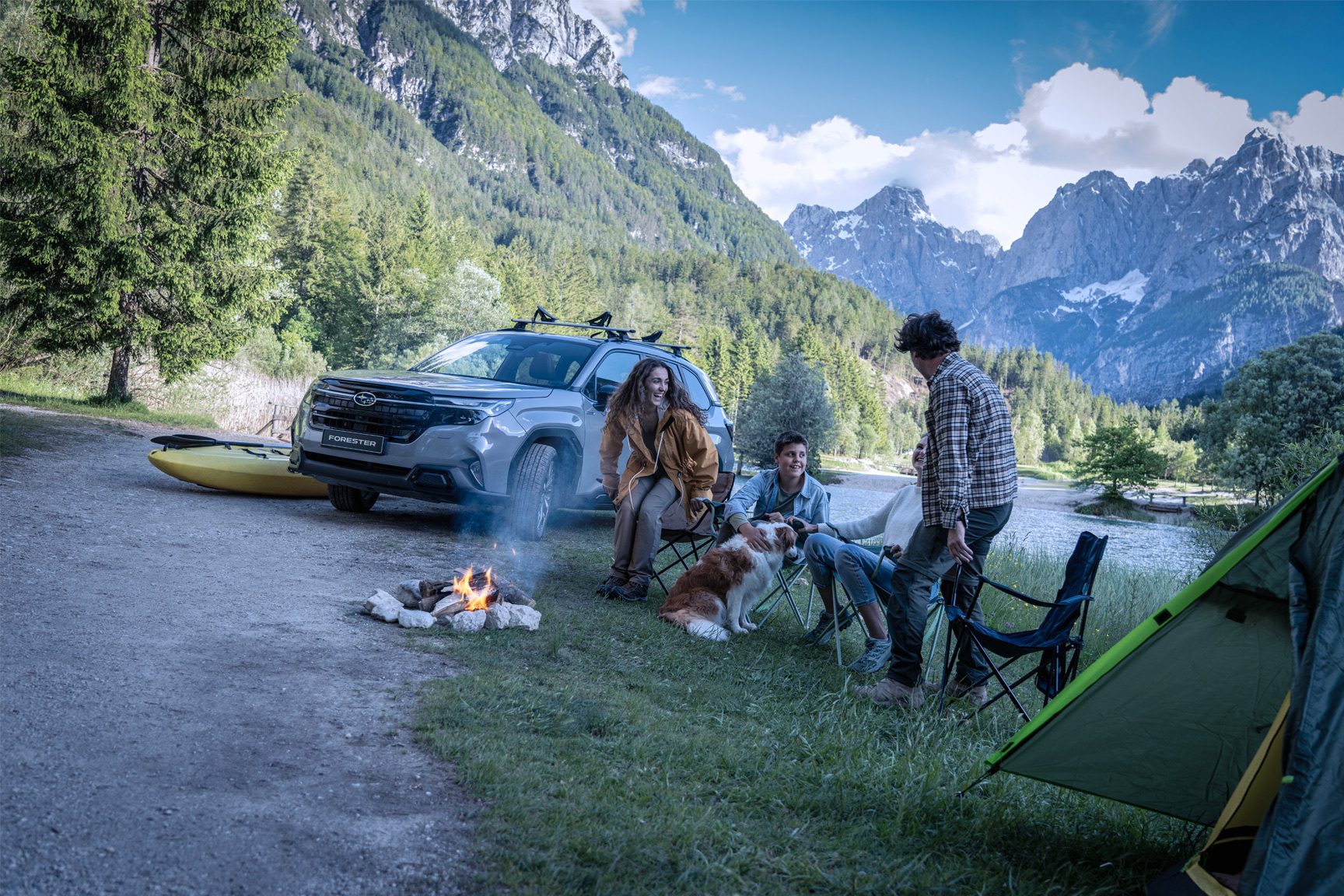 Group camping by a campfire with a Subaru Forester, kayak, dog, and mountains.