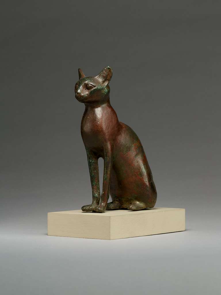 Ancient Egyptian bronze statue of a seated cat with green and reddish patina, looking left.