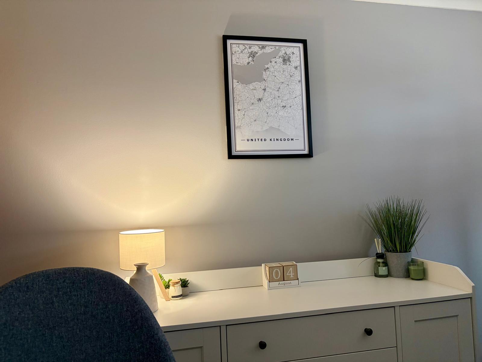 Minimalist home office with a framed UK map, a warm lamp, a perpetual calendar, and greenery on a light gray desk.