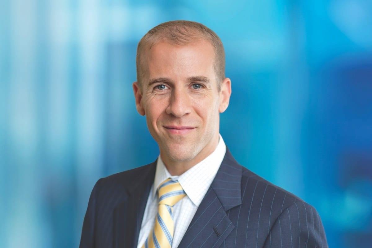 Headshot of a smiling man in a pinstripe suit and yellow striped tie against a blue background.
