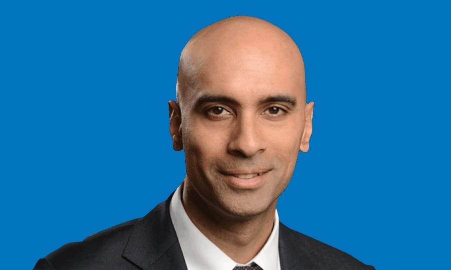 A professional headshot of a bald man with dark eyes and a slight smile against a blue background.