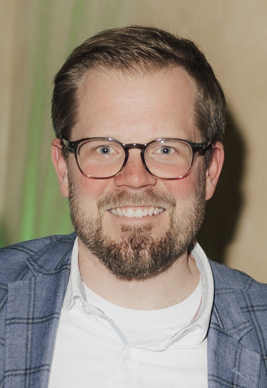 Smiling man with glasses and a beard in a blue plaid jacket.