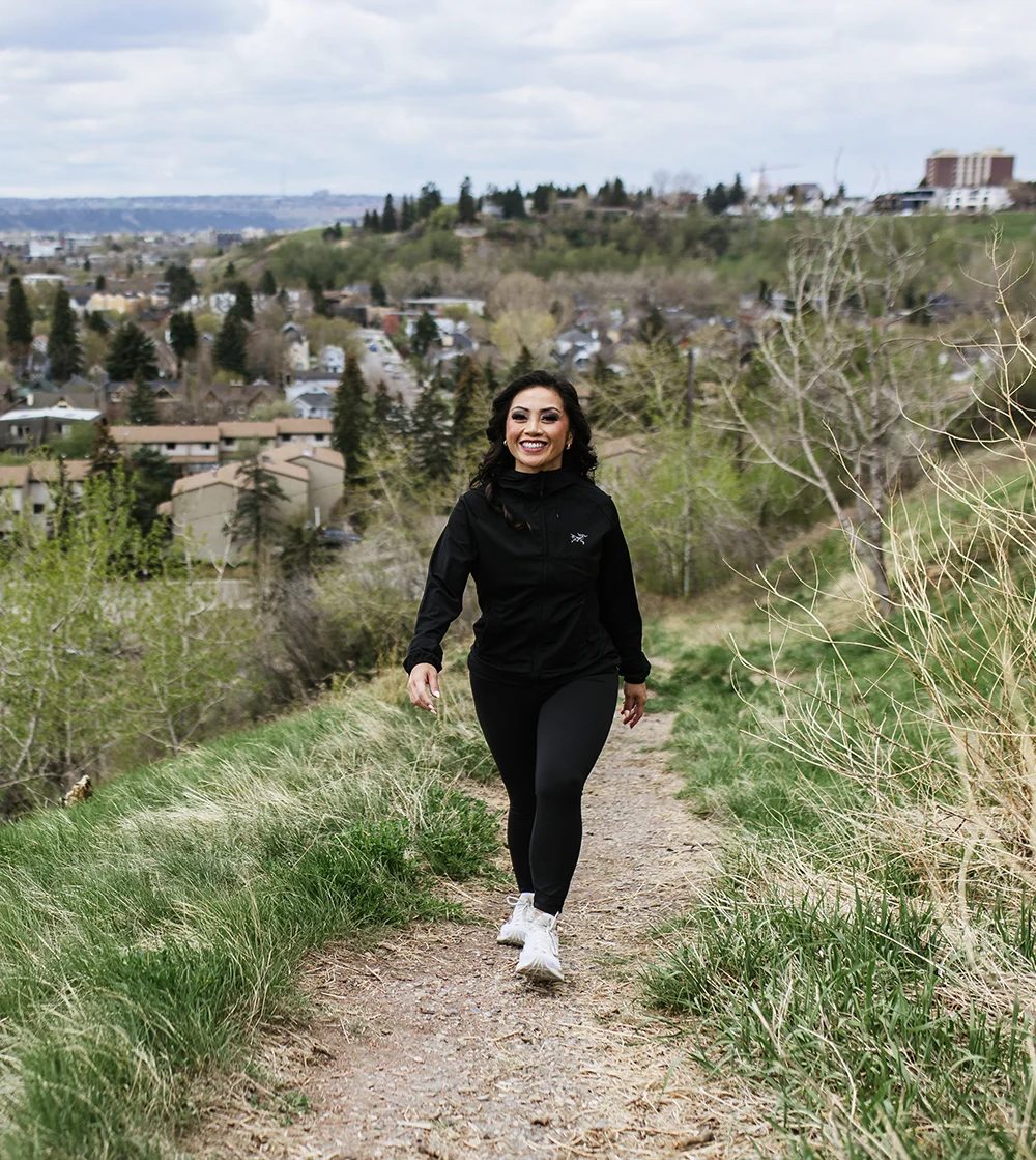 A smiling woman in black activewear walks on a dirt path outdoors, with a town in the background.