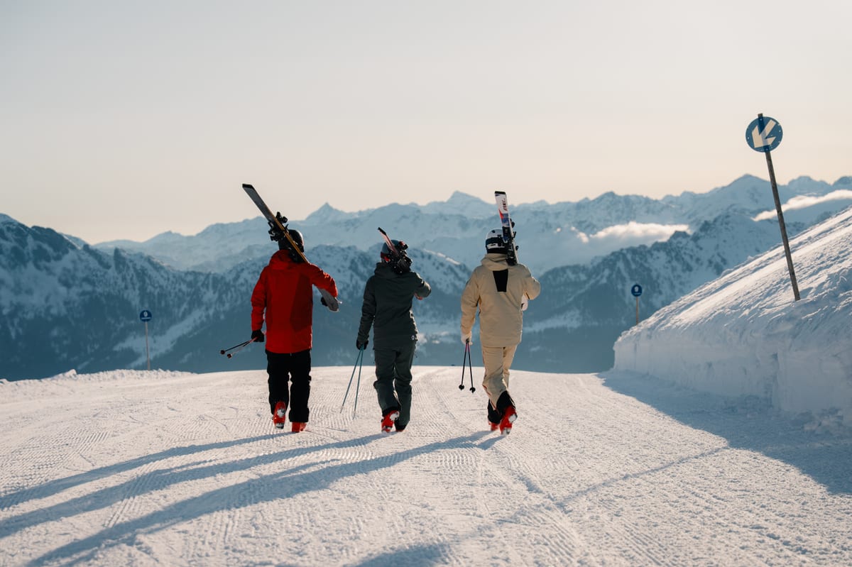 Three skiers walking up a snowy mountain path with skis on their shoulders, mountains in the background.