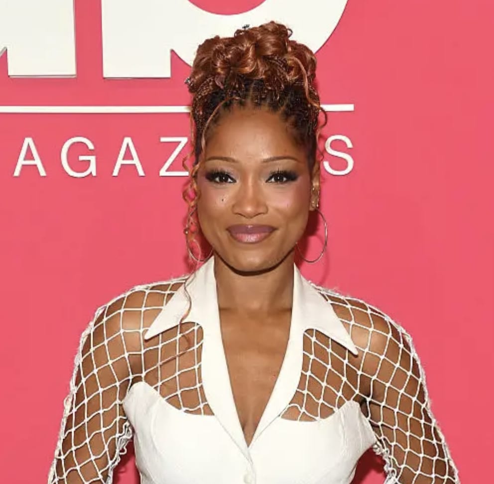 Keke Palmer smiling, wearing a white mesh-detailed top and hoop earrings against a pink background.