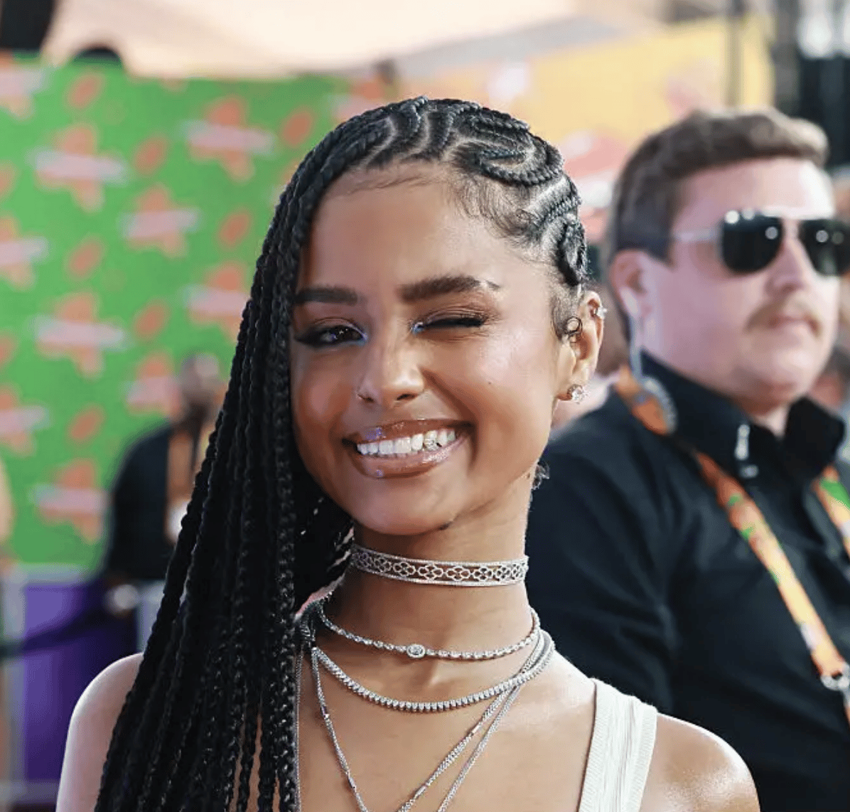 Young woman with intricate braided hair winking and smiling, wearing layered necklaces, at an event.