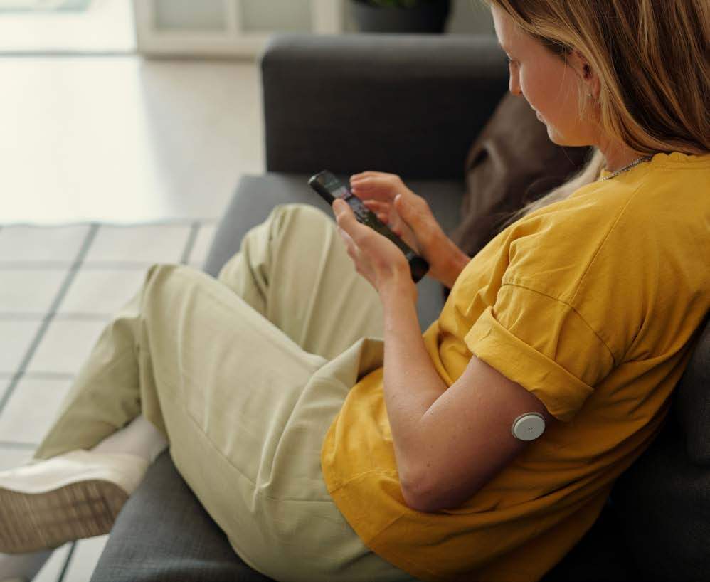 A woman relaxing on a couch, using a TV remote and wearing a continuous glucose monitor (CGM) on her arm.