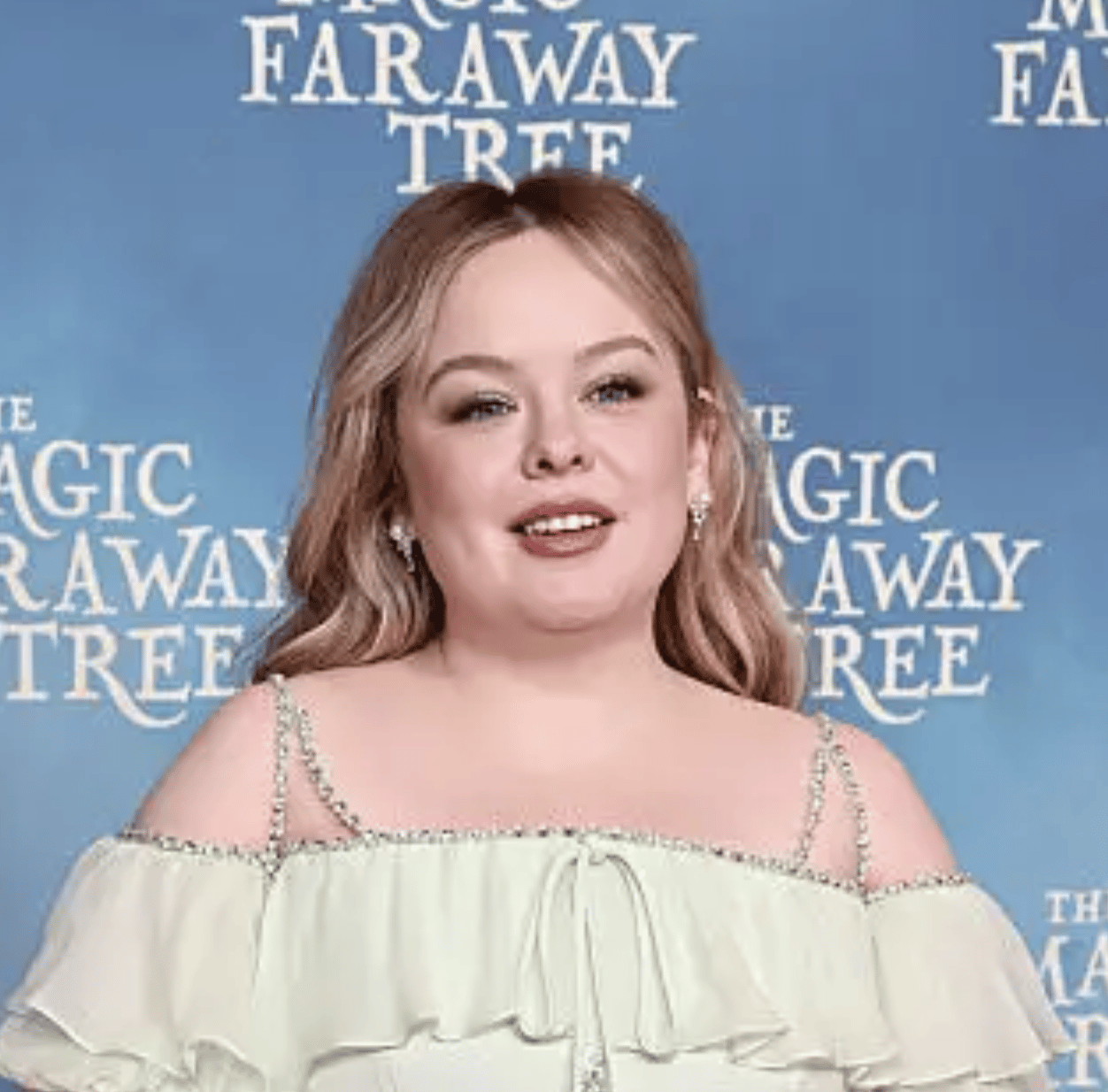 Nicola Coughlan smiles in a light green off-the-shoulder dress against a blue Faraway Tree backdrop.
