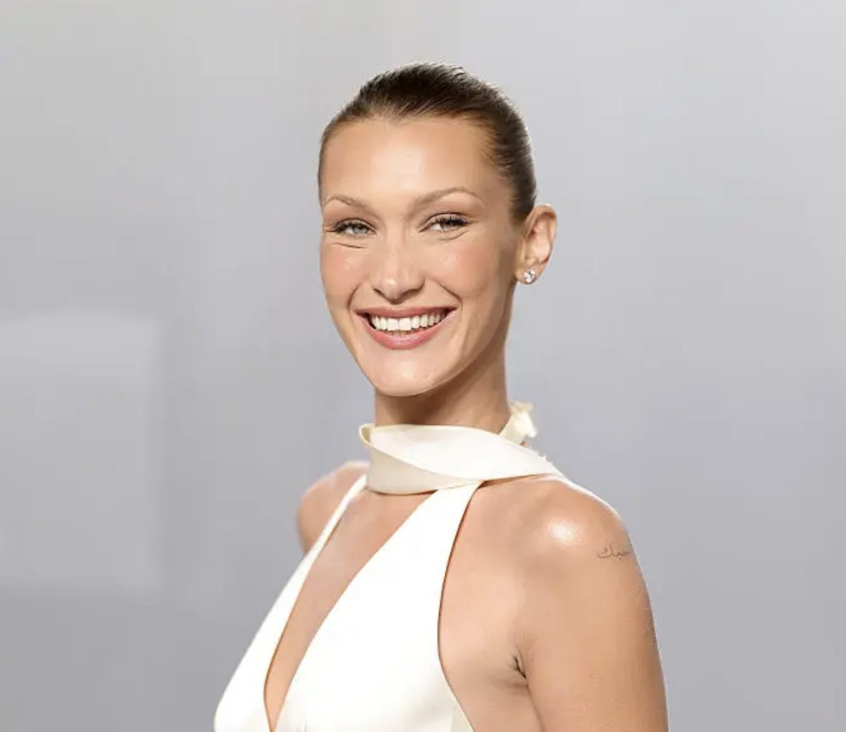 Bella Hadid smiles brightly, wearing a white halter-neck dress against a plain background.