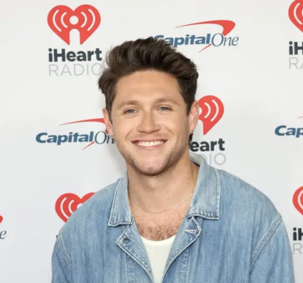 Niall Horan, smiling, wearing a denim jacket over a white shirt, against a white background with logos.