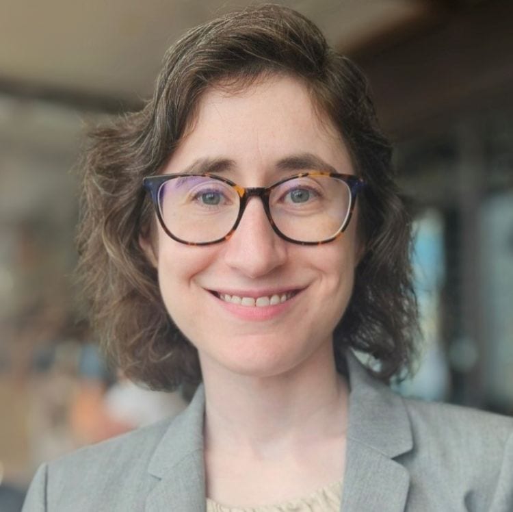 Smiling woman with short brown hair, glasses, and a grey blazer, looking at the camera.