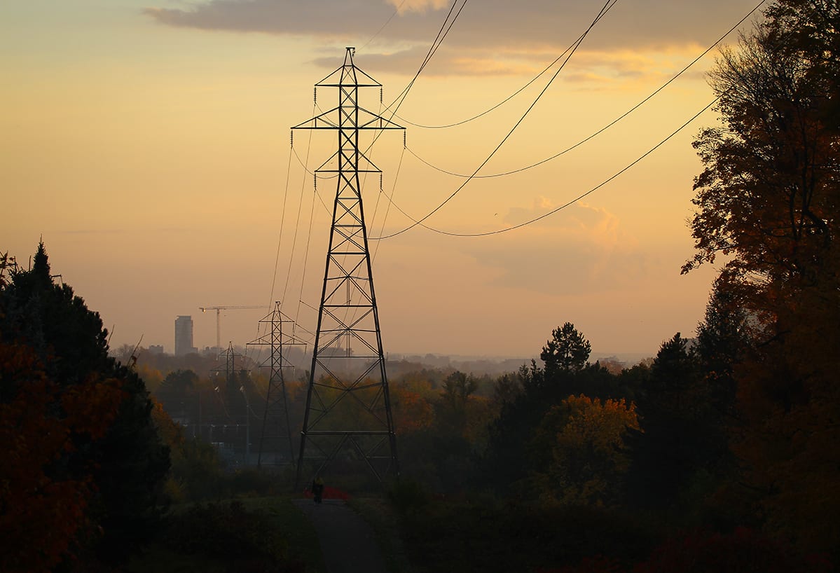 Power lines and towers silhouette against a hazy sunset sky over trees and distant buildings in Waterloo Region.