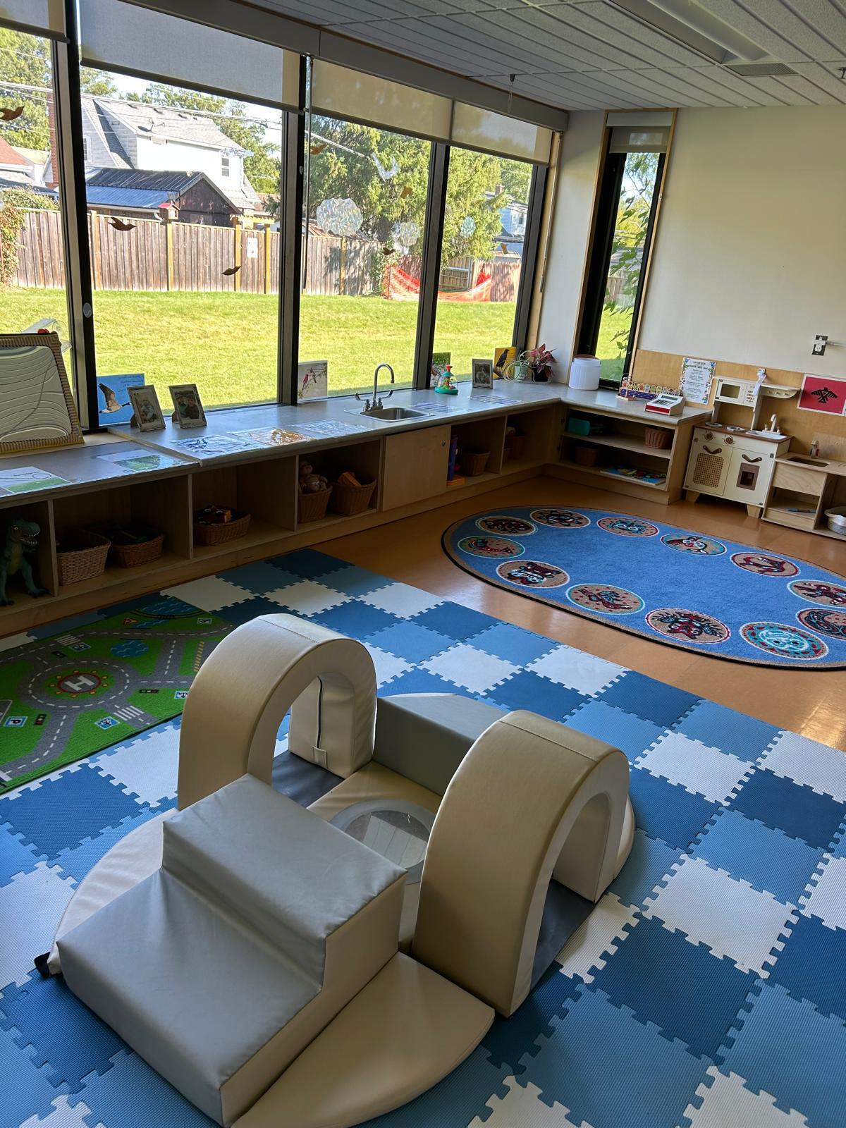 Child care centre play stations, with a soft climbing toy, play kitchen and storage shelves with toys. The space is surrounded by large, bright windows, looking out onto a lawn. 