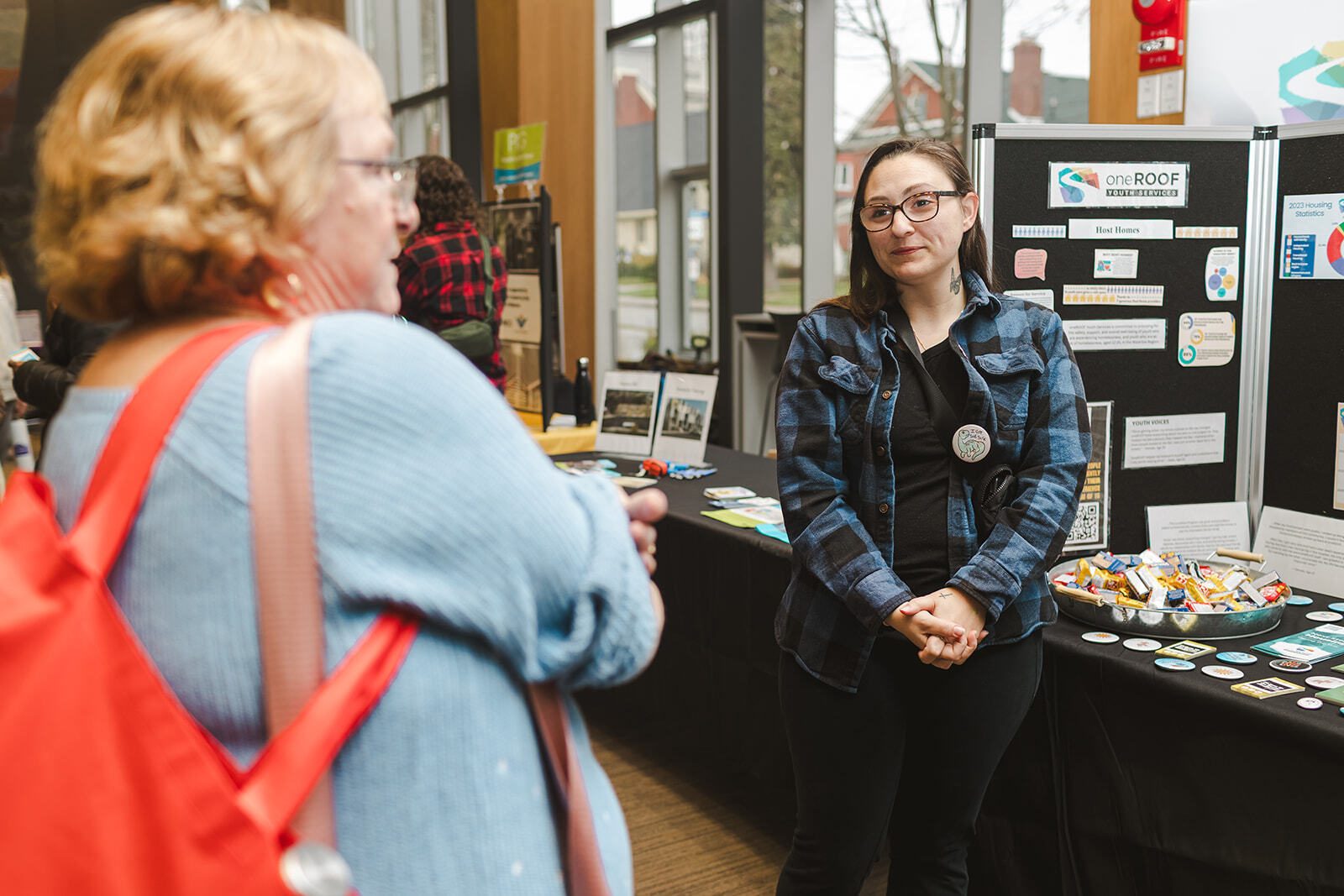 An individual stands at an information booth at an event at the Kitchener Public Library. They have a display set up on a table. The logo for housing services non-profit OneRoof is posted on the display. A person is out-of-focus in the foreground speaking with the individual at the booth. 