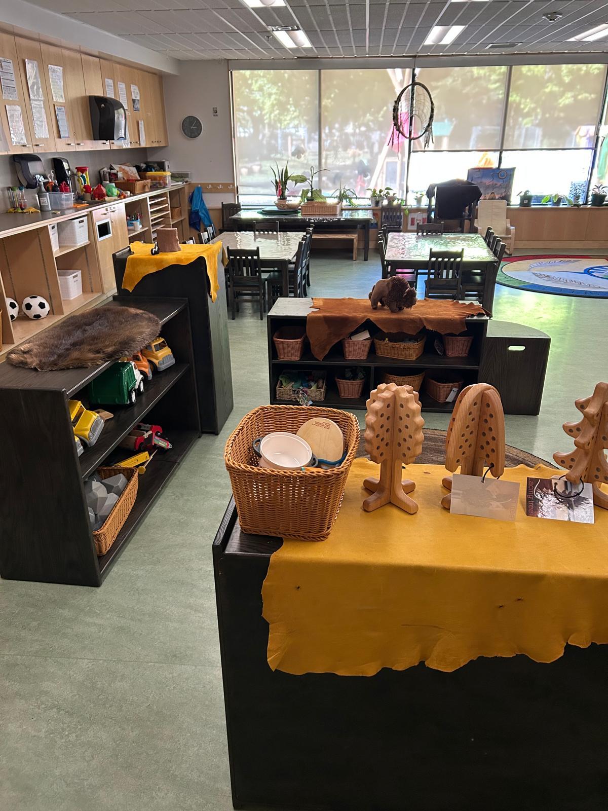 Child care centre space with shelves with toys, including wooden trees in the foreground, and 2 child-height tables with chairs in the background.   The room has furniture, nature-inspired toys, and elements like fur and leather.