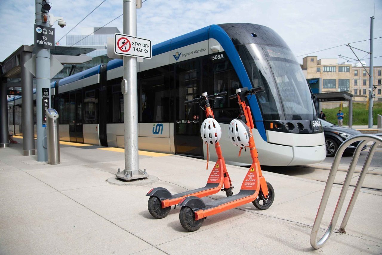 Two neuron scooters parked on the sidewalk. Behind them is the ION train at a Cambridge stop.