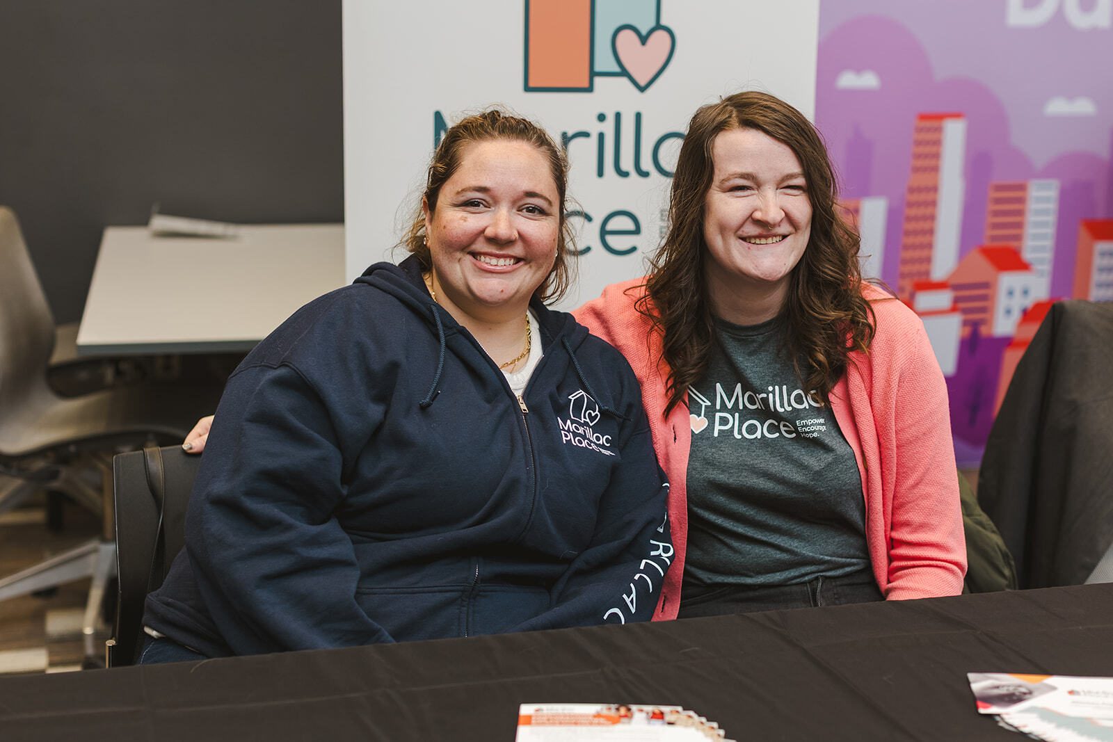 Two people are seated at an information booth. They are wearing t-shirts with a logo from the non-profit Marillac Place. A stand-up banner is behind them with the same logo. Both people are smiling. 