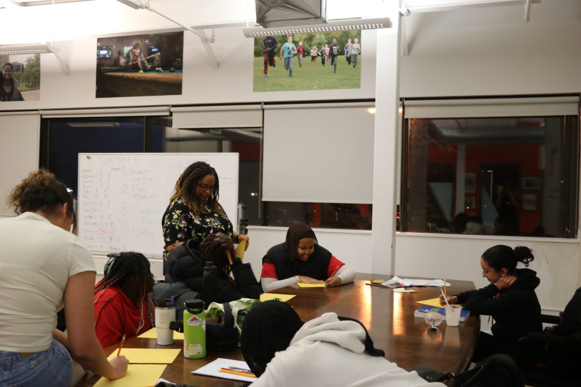 A diverse group of young people writing at a large table indoors, with an adult supervising.