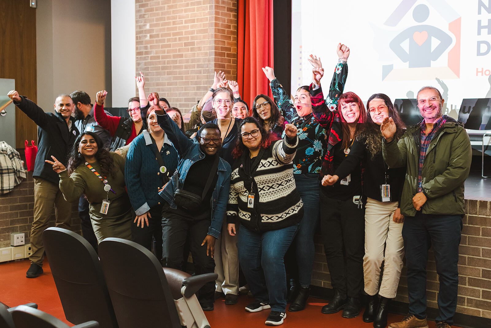 A group of people celebrating National Housing Day at an event hosted by the Region. The people are gathered at the front of an auditorium-style room with a stage behind them. The people are from diverse groups and are all happy. Many people are smiling while pushing their fist into the air.  