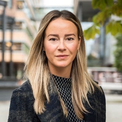 A blonde woman in a dark coat and patterned top looks directly at the camera.