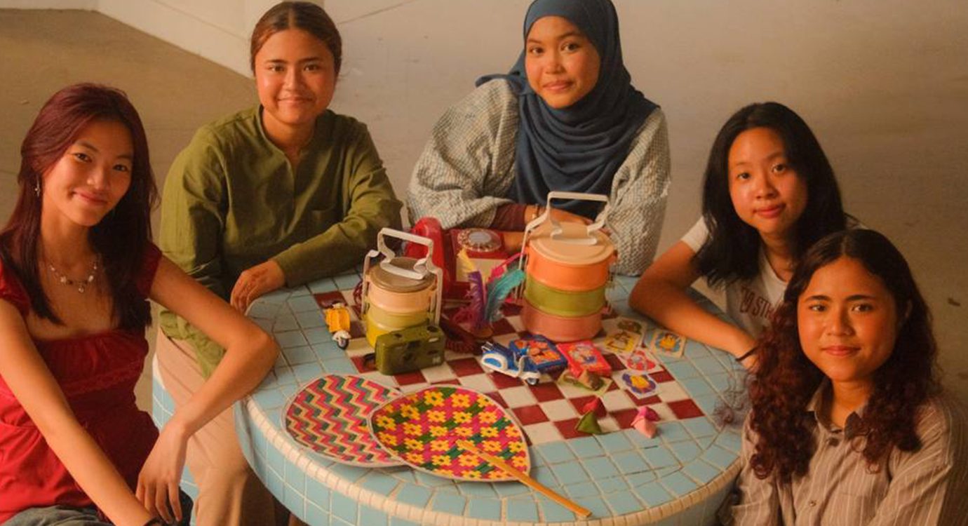 Five young women around a table with patterned tiffin carriers and a fan.