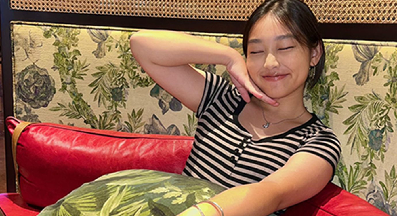 Young woman with eyes closed, smiling, hand on chin, sitting on a botanical print chair.