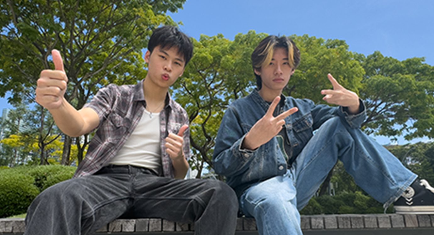 Two young men pose outdoors. One gives thumbs up; the other makes a peace sign and points.