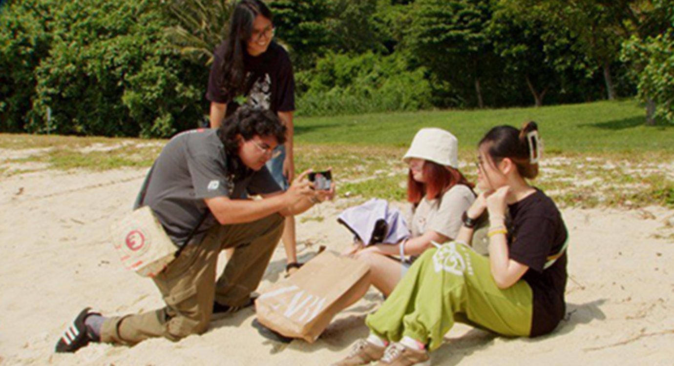 Four friends outdoors: one kneels taking a photo of two sitting on sand, another stands.