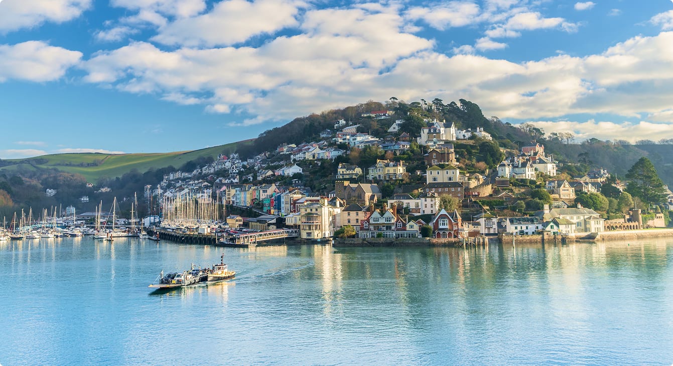 Colorful town on a hillside by a river with boats & ferry, under a blue, cloudy sky.