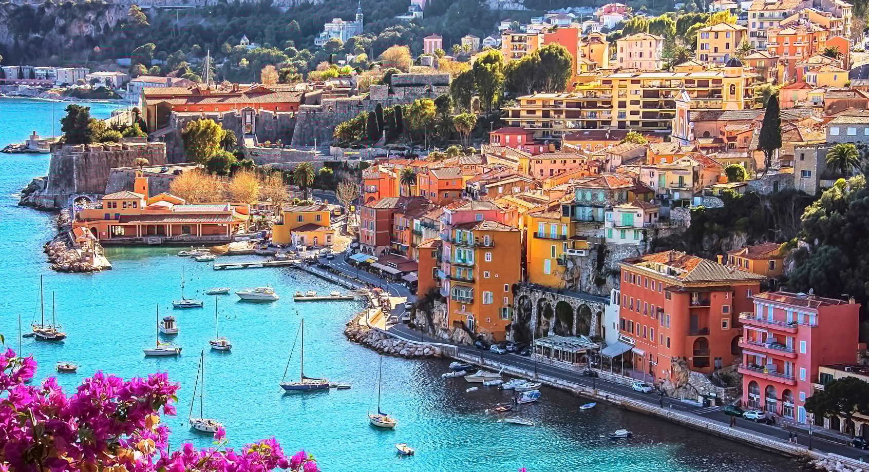 Vibrant Mediterranean town with colorful buildings, a turquoise bay with boats, and purple bougainvillea.