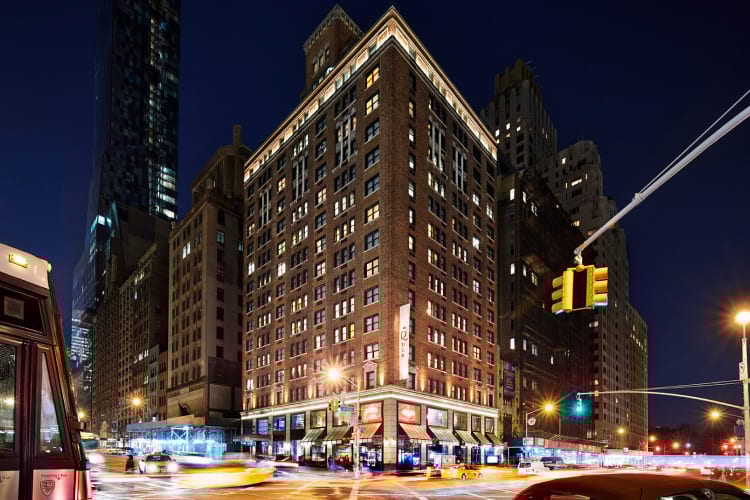Night view of a vibrant city street corner with a lit-up brick building and car light trails.
