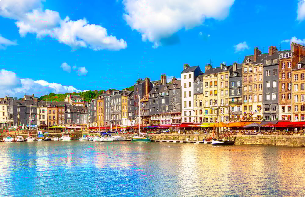 Colorful historic buildings line a bustling harbor with boats under a bright blue sky in Honfleur.