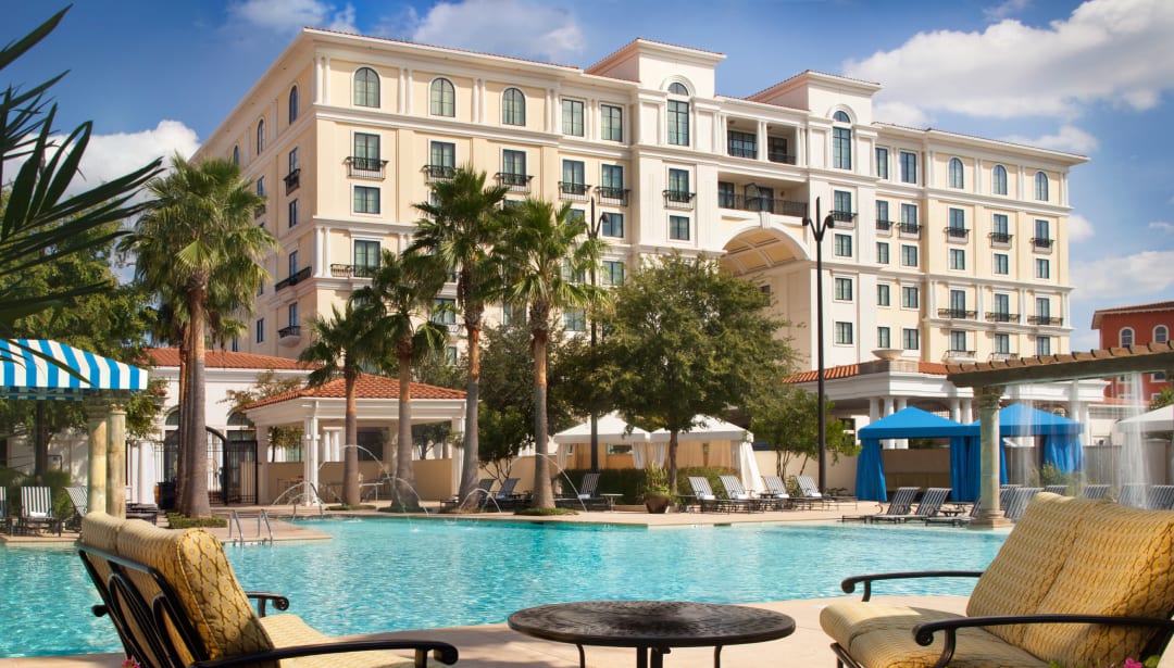 Resort hotel with a large swimming pool, palm trees, and lounge chairs under a blue sky.