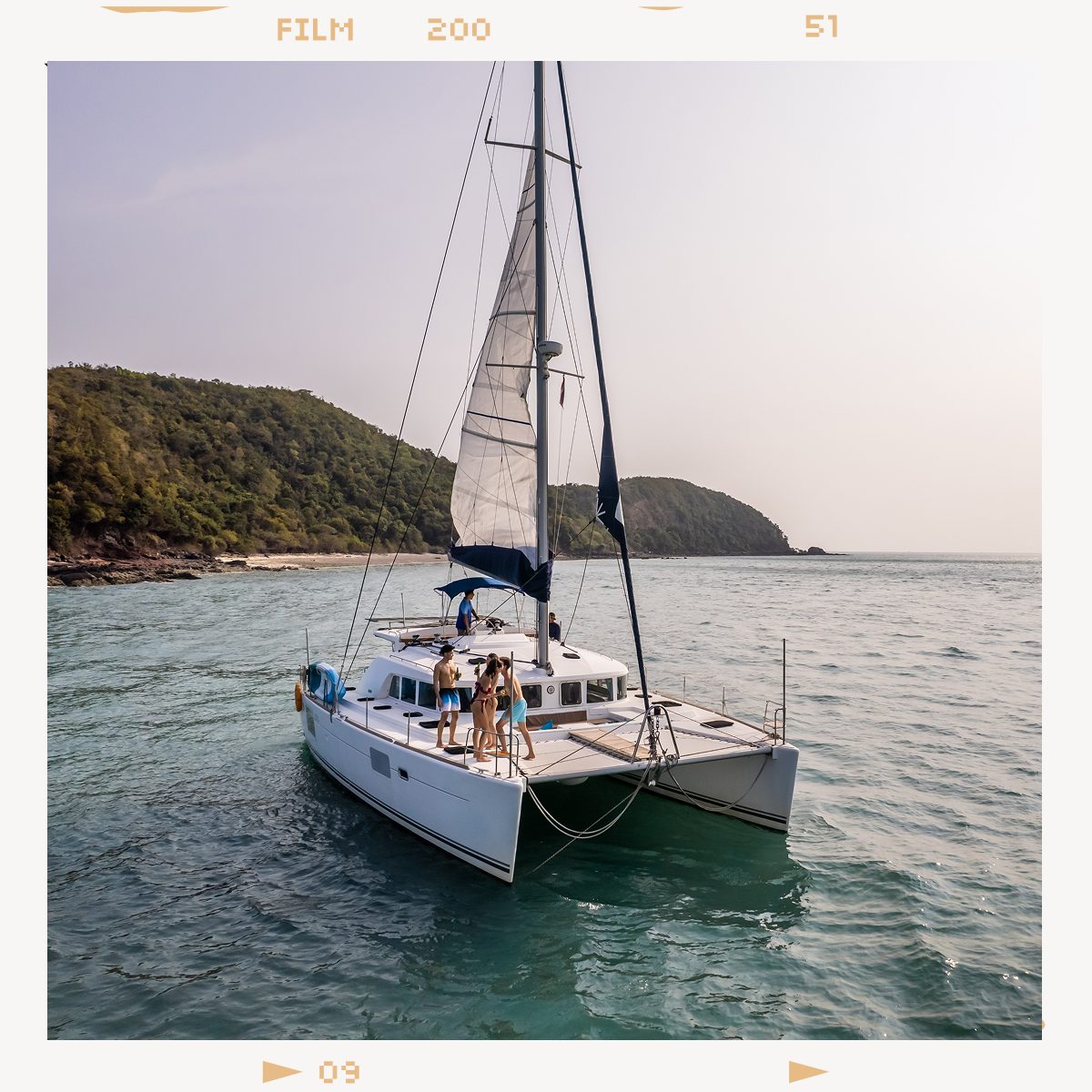 A white catamaran sailboat with people on deck, sailing on clear blue water near a lush island.