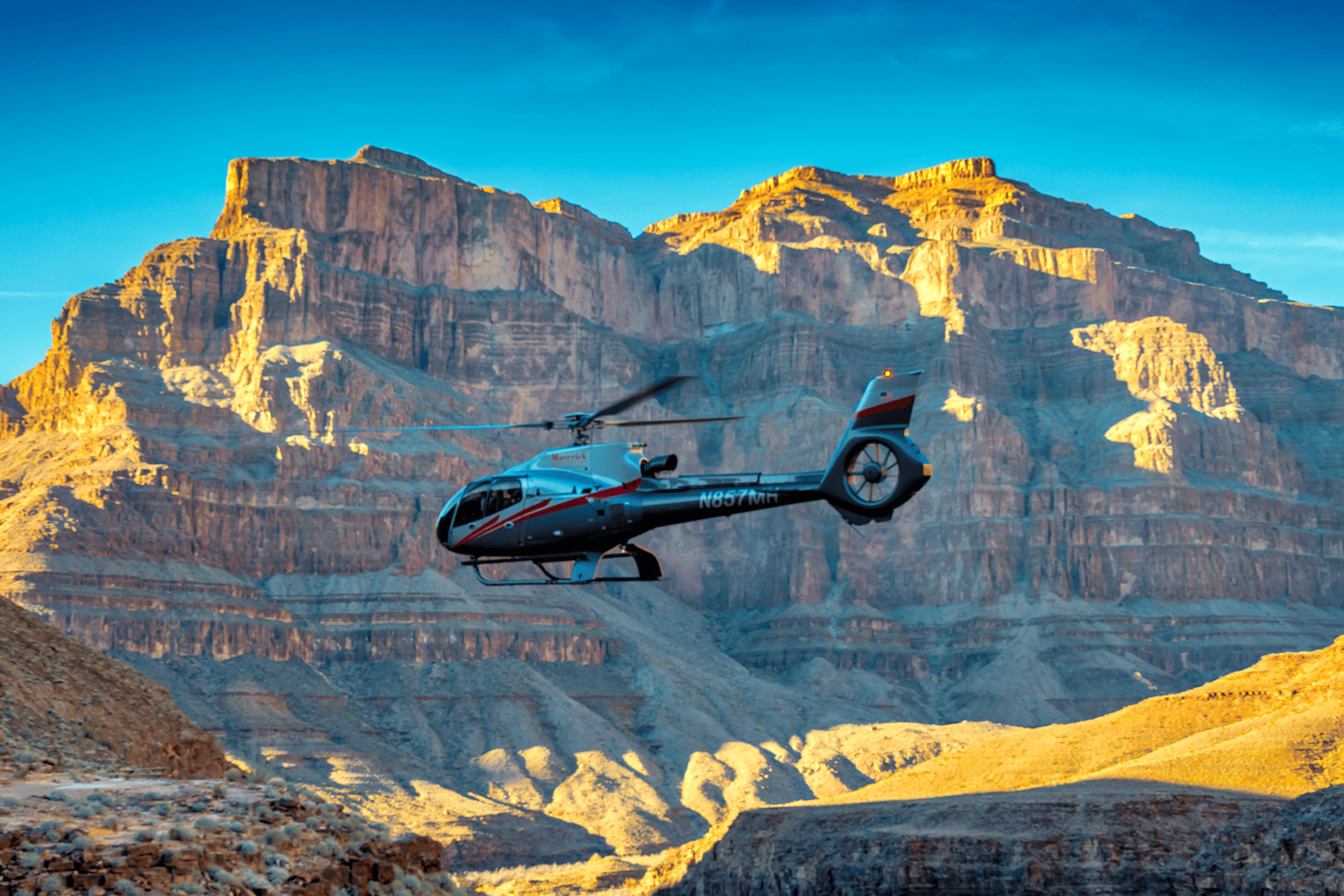 A grey helicopter flies over a vast, sunlit canyon with towering rock formations.