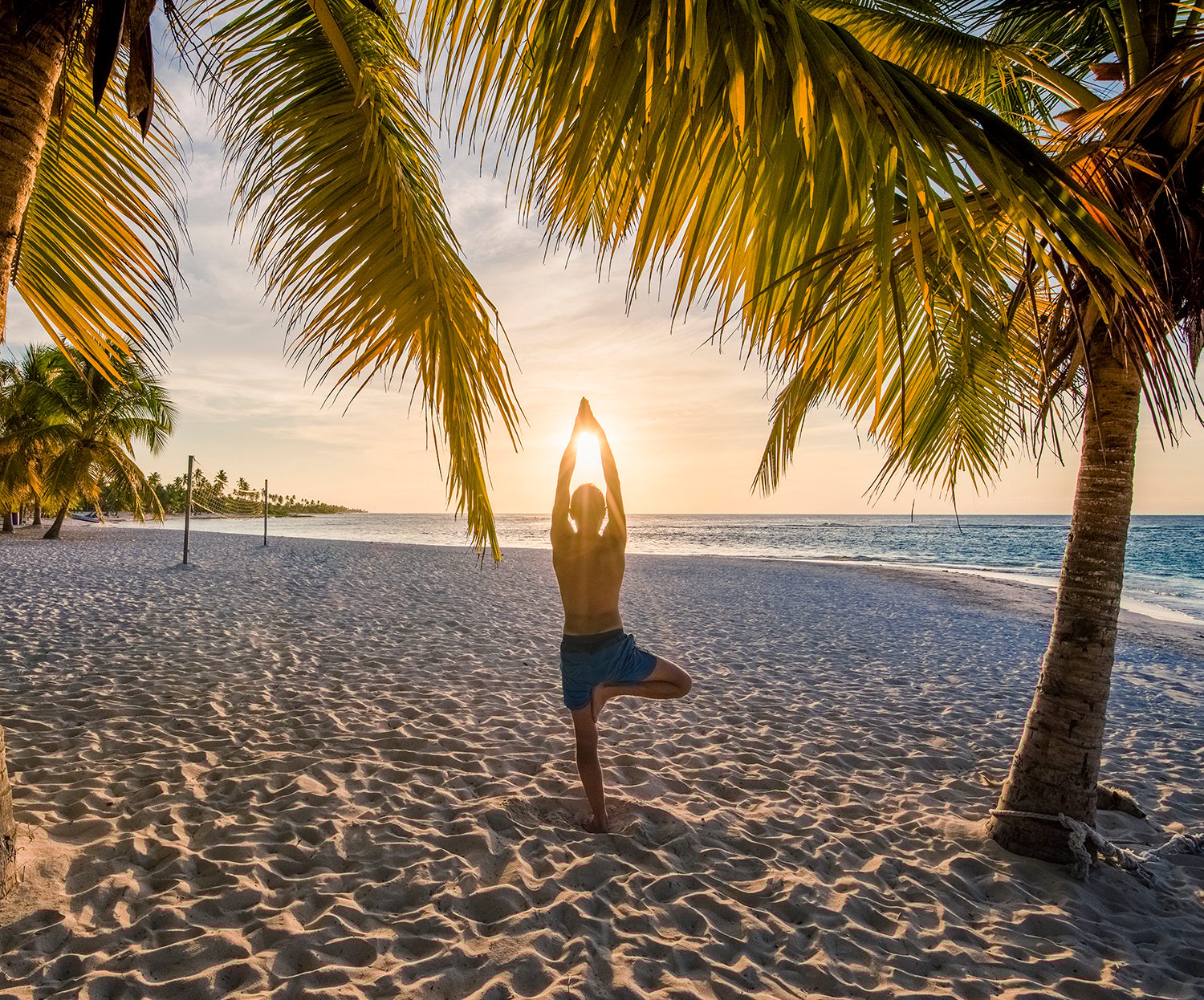Man doing tree pose yoga on a sandy beach at sunset, framed by palm trees.