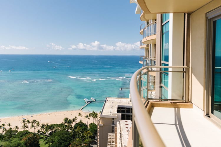 High-angle view from a balcony showing a tropical beach, clear blue ocean with boats, and a pier.