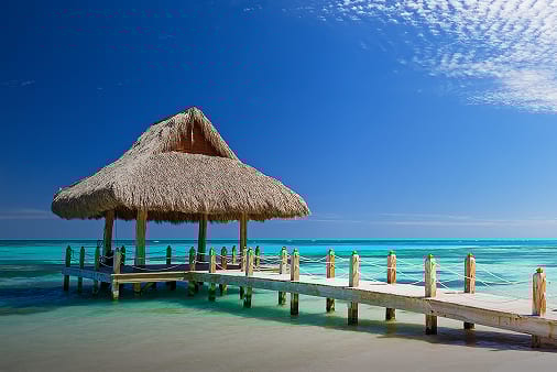 A serene thatched gazebo stands on a wooden pier over crystal-clear turquoise waters under a bright blue sky.
