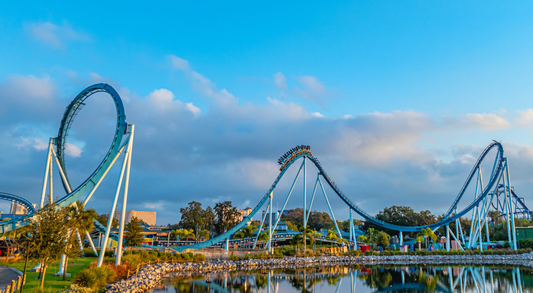 Blue roller coaster with loops and riders reflected in water under a blue sky.