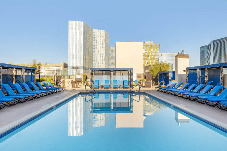 Rooftop swimming pool with blue lounge chairs, cabanas, and city skyscrapers under a clear sky.