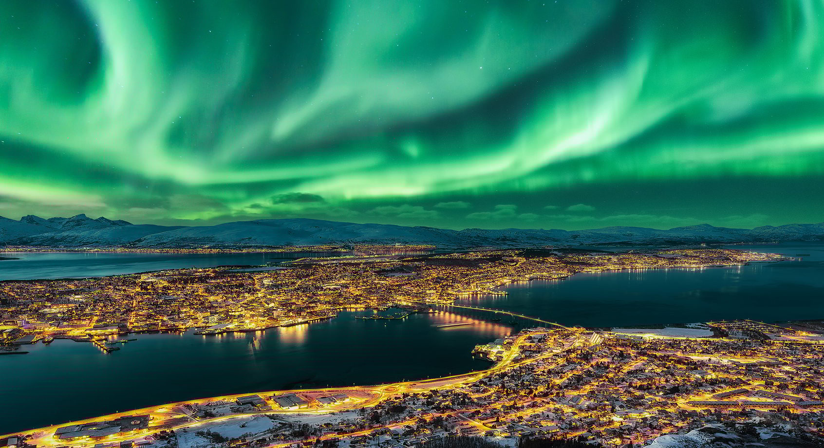 Aurora Borealis over a brightly lit city by a fjord, with snow-capped mountains.
