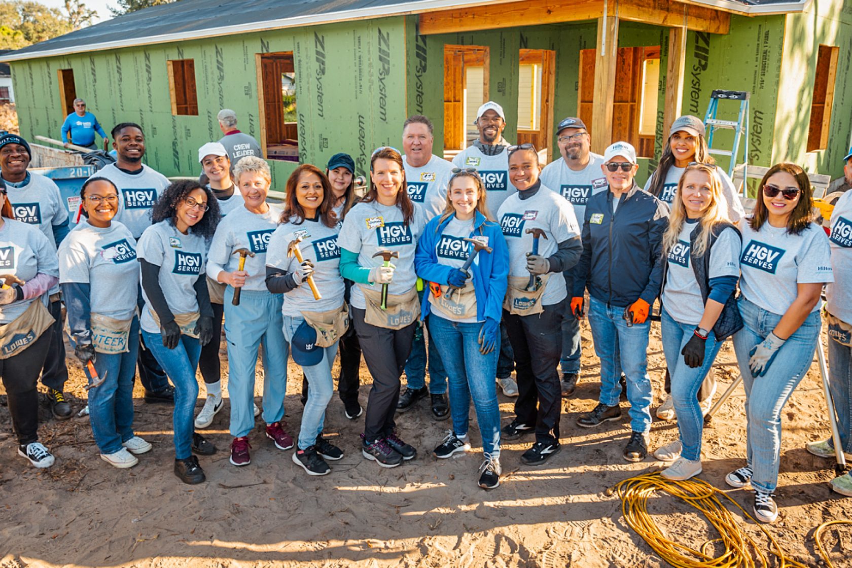 Smiling volunteers from "HGV SERVES" pose with hammers in front of a house under construction.