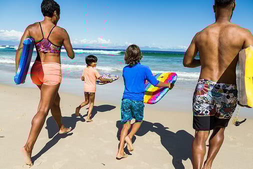 Family running with bodyboards towards the ocean on a sunny beach.
