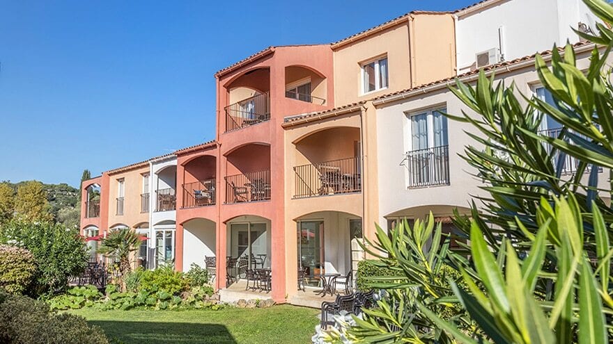 Colorful resort apartments with balconies and ground-floor patios, surrounded by a lush green garden.