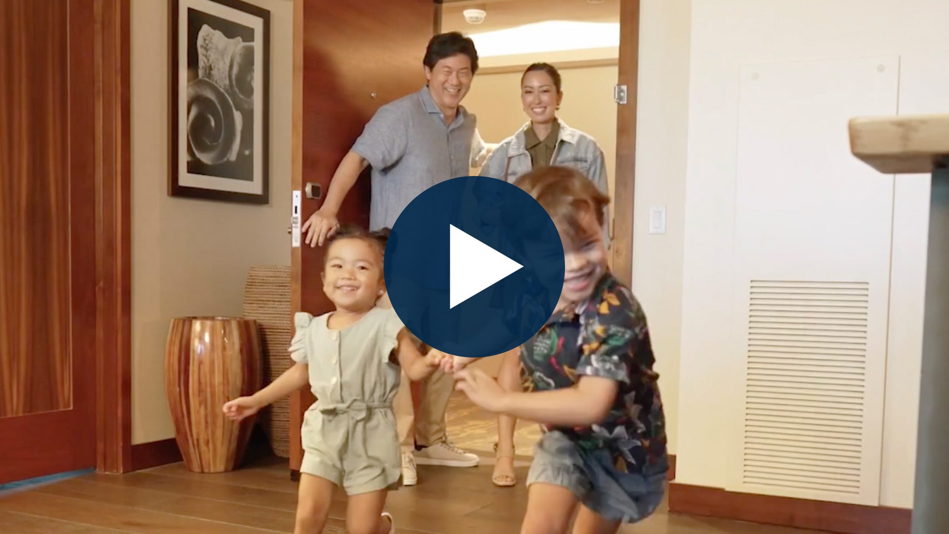 A smiling family with two adults and two young children joyfully entering a room, captured from a video.