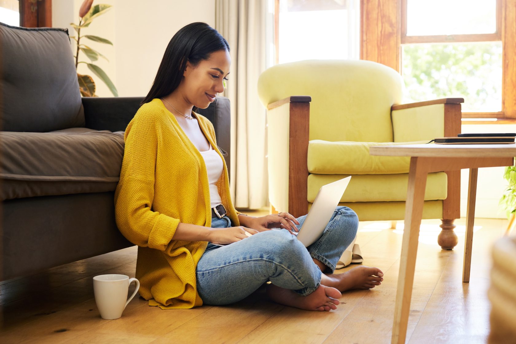 A smiling woman sits on a wooden floor, leaning against a sofa, working on her laptop.